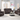 A living room setup featuring a reclining armchair, loveseat, and sofa in rich brown leather with nailhead trim, situated around a round coffee table. The room has large windows letting in natural light and modern art pieces on the walls.