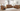 A cozy living room featuring a brown leather reclining sofa set including a reclining loveseat with cupholders, a reclining armchair, and a comfortable sofa. The furniture is arranged around a light-colored rug, with large windows providing natural light.